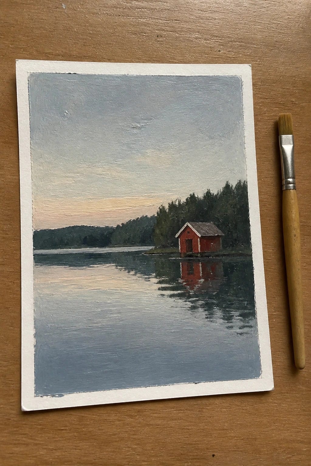 Red cabin reflected in calm lake at dusk beside paintbrush on wooden table