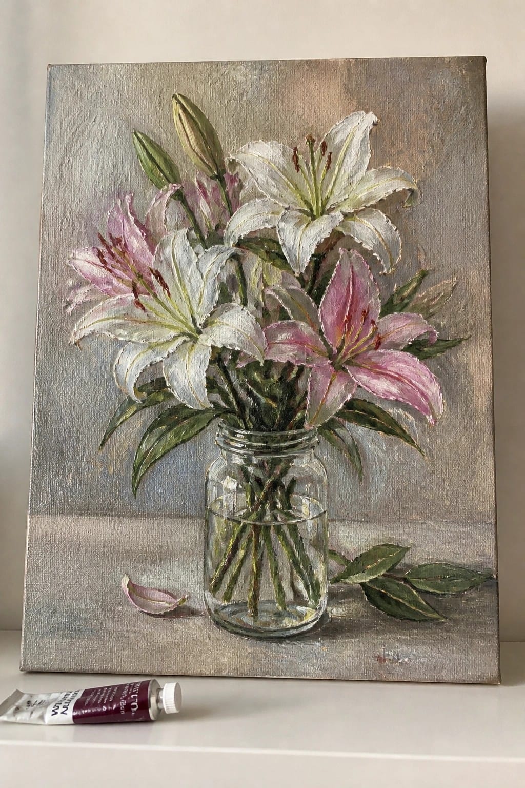 Oil painting of pink and white lilies in a glass jar with visible stems in water on a gray surface, with a paint tube nearby.
