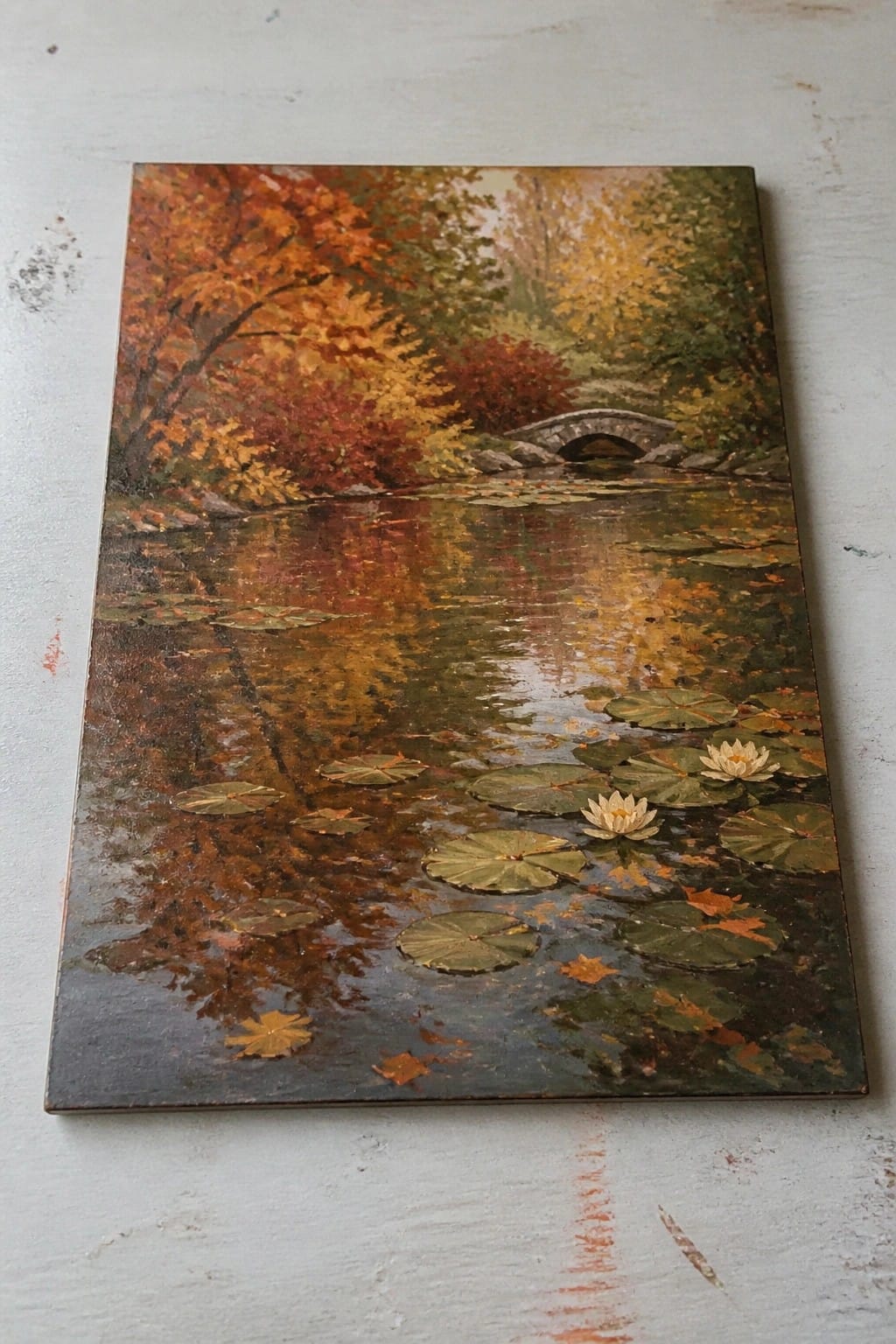 Autumn pond with lily pads, white flowers, red-orange trees, and stone bridge reflections.