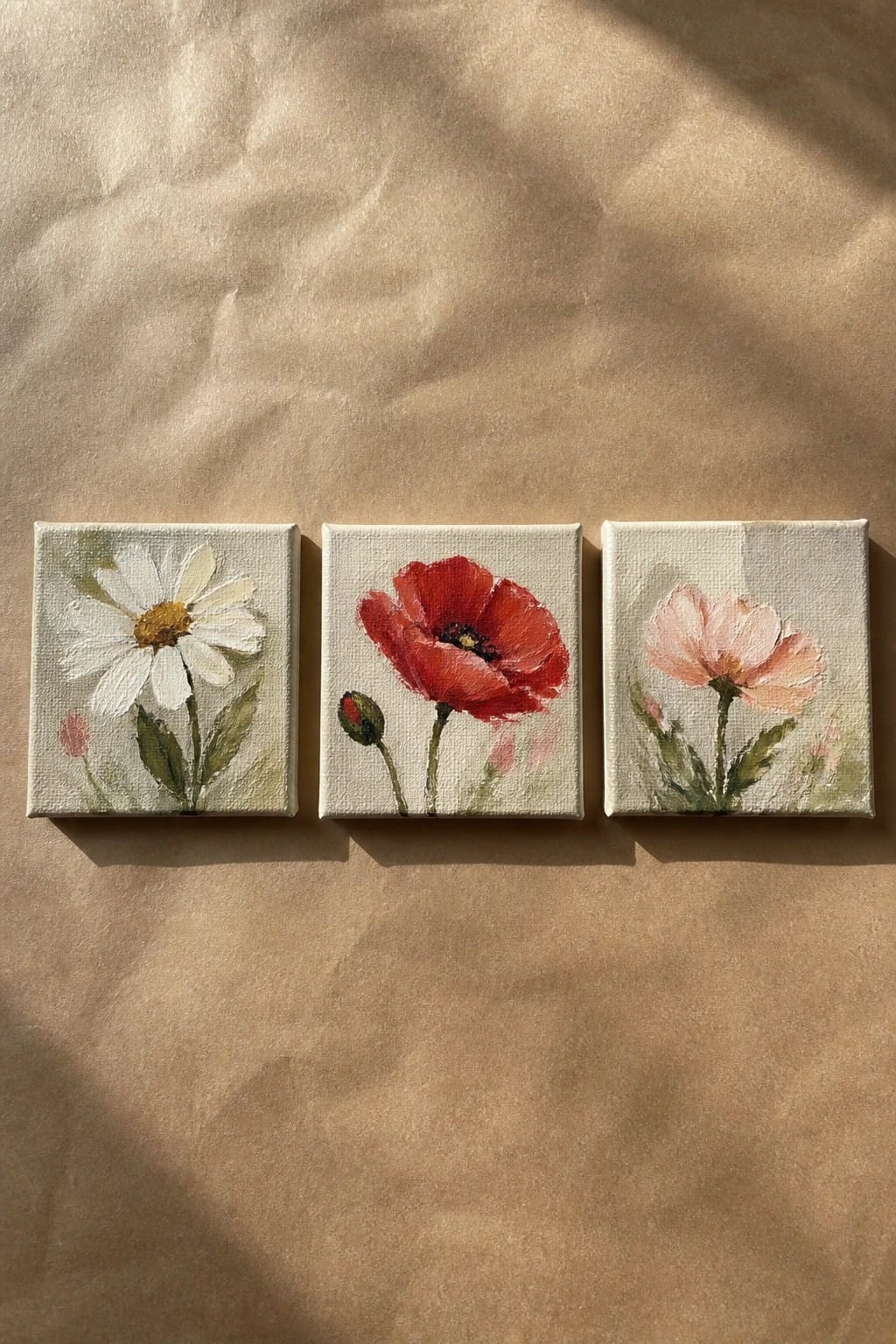 Three square canvases with white daisy, red poppy, and pink flower on brown paper