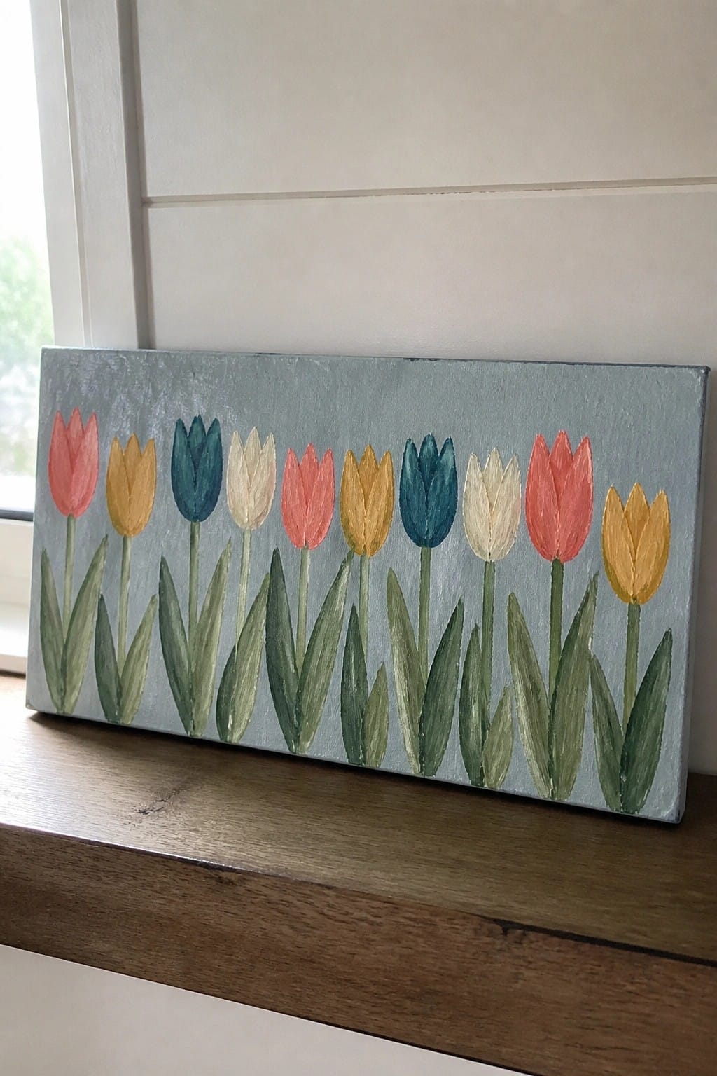 Colorful tulips painted on light blue canvas resting on wooden windowsill.