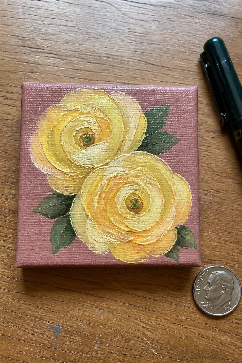 Two yellow roses painted on small pink canvas beside green pen and dime.
