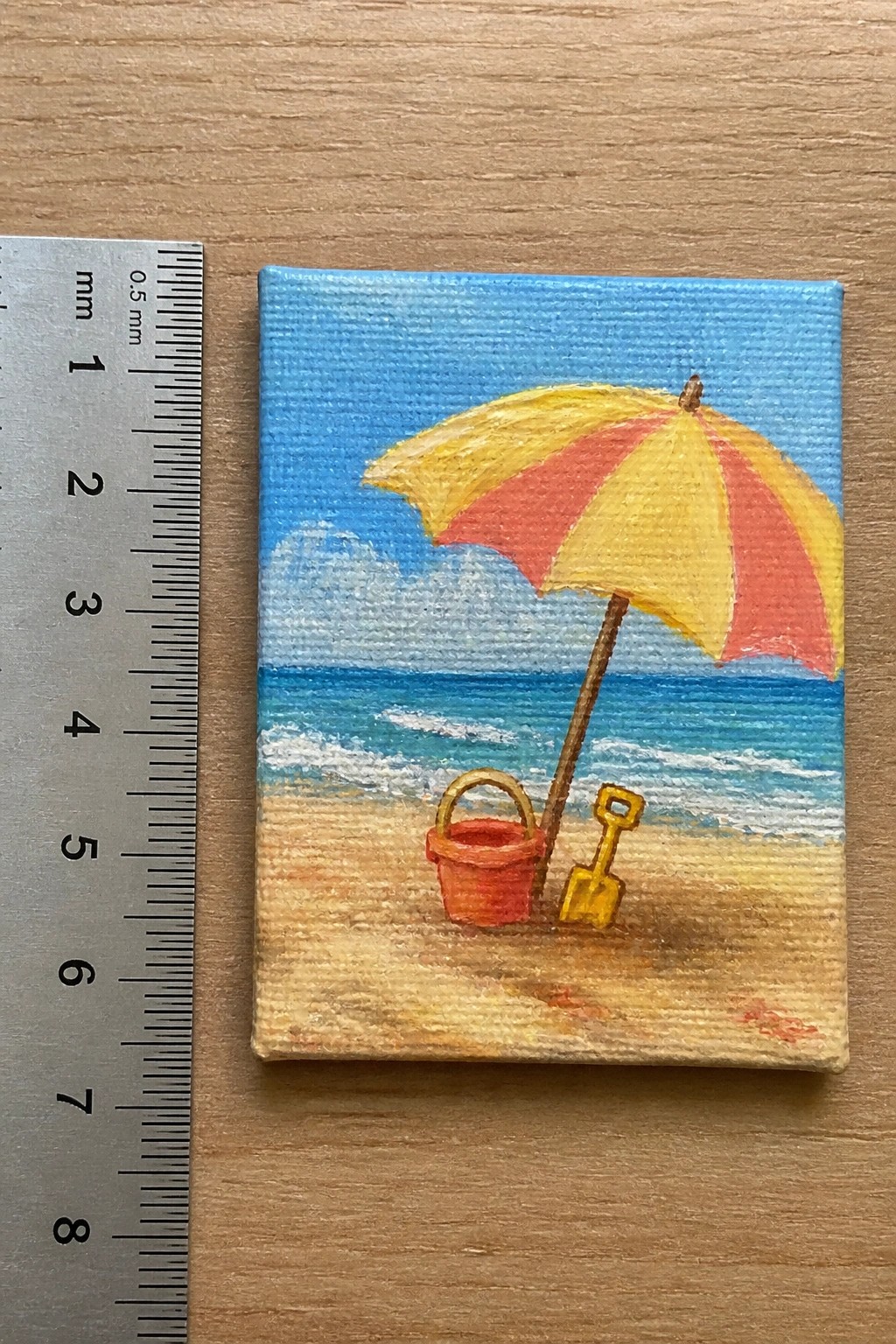 Miniature beach painting with striped umbrella, red bucket, yellow shovel beside ruler