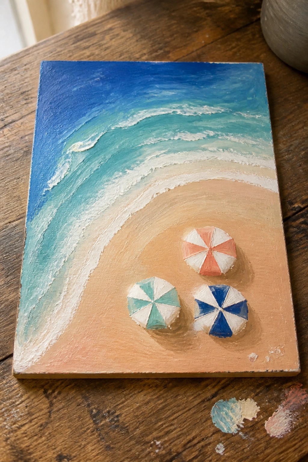 A small oil painting of a beach with blue and turquoise waves, light sand, and three colorful umbrellas resting on the shore.