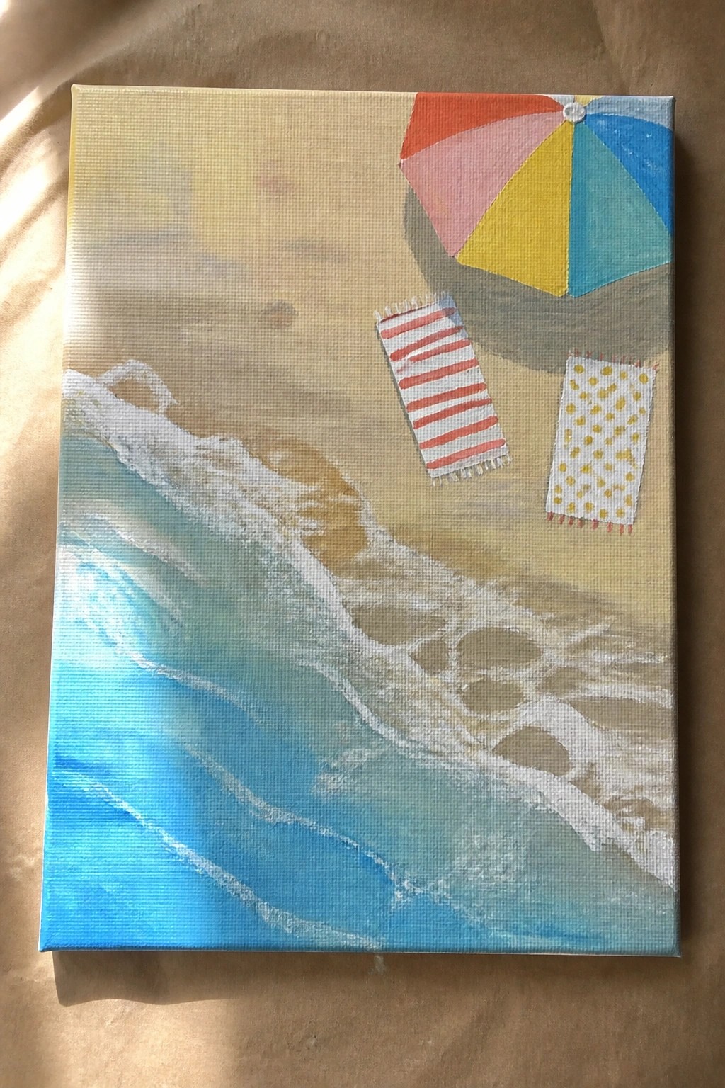 Colorful beach umbrella over striped and dotted towels beside blue ocean waves on sand.