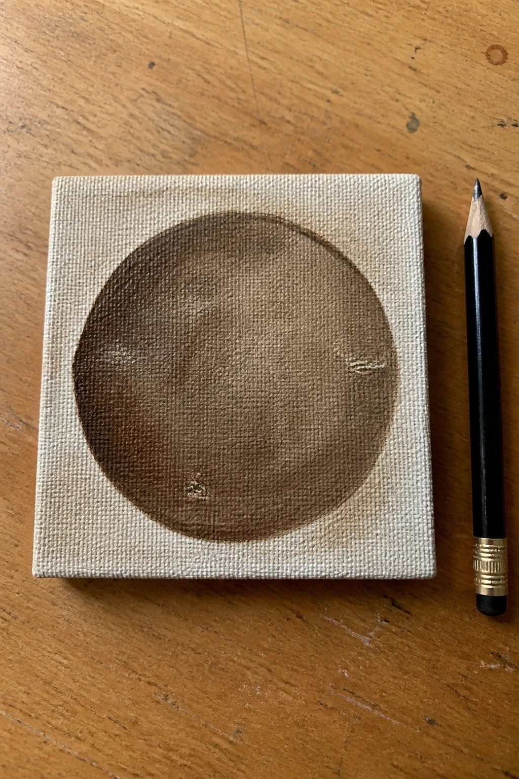 Small canvas with textured brown moon painting beside black pencil on wood.