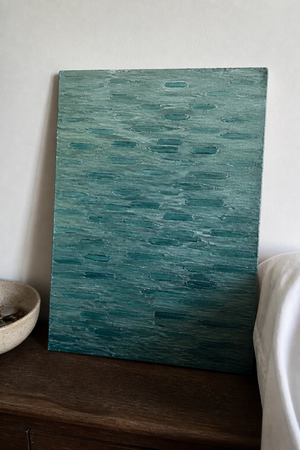 Textured teal-green abstract painting with horizontal strokes on wood beside bowl and cloth