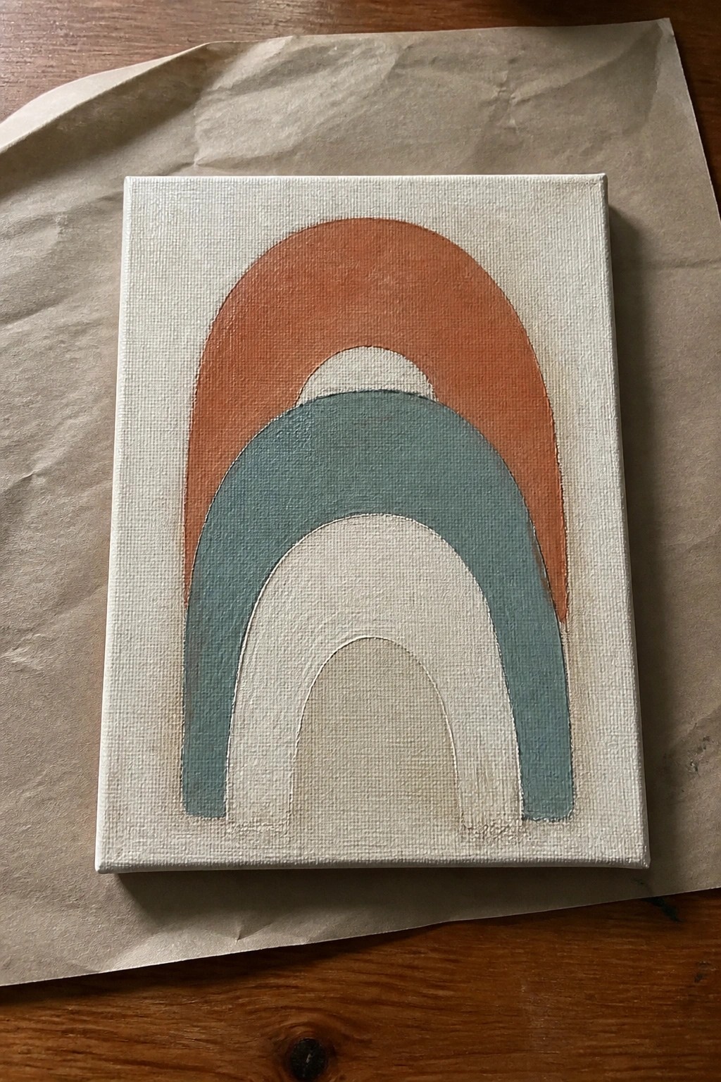 Canvas with stacked rainbow arches in orange, teal, and white on brown paper over wood