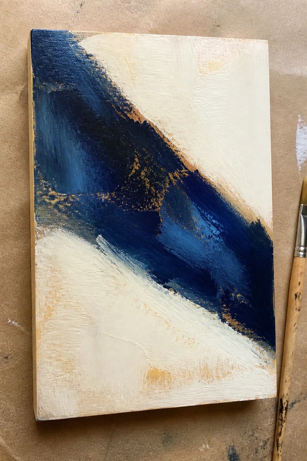Abstract painting with blue, white, and gold brushstrokes beside paintbrush.