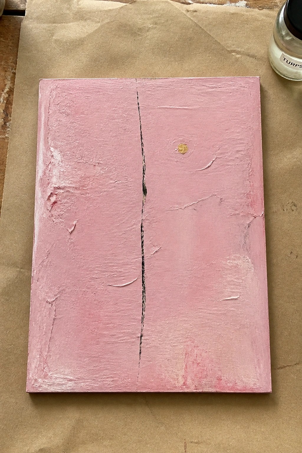 Pink textured painting with black vertical crack, gold dot, on brown paper beside turpentine jar