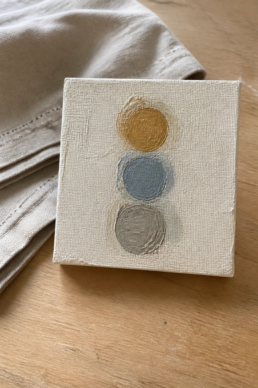 Small white canvas with gold, blue, and gray circles beside beige fabric on wood