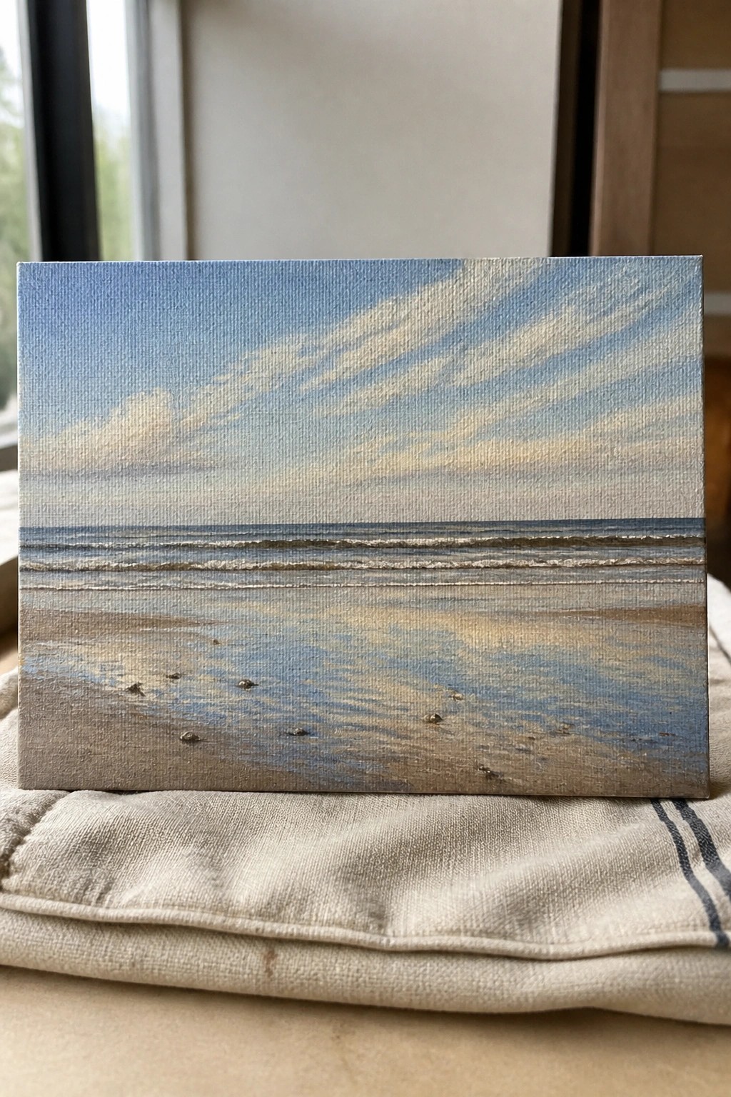 Small canvas beach painting with blue sky and ocean on striped fabric.