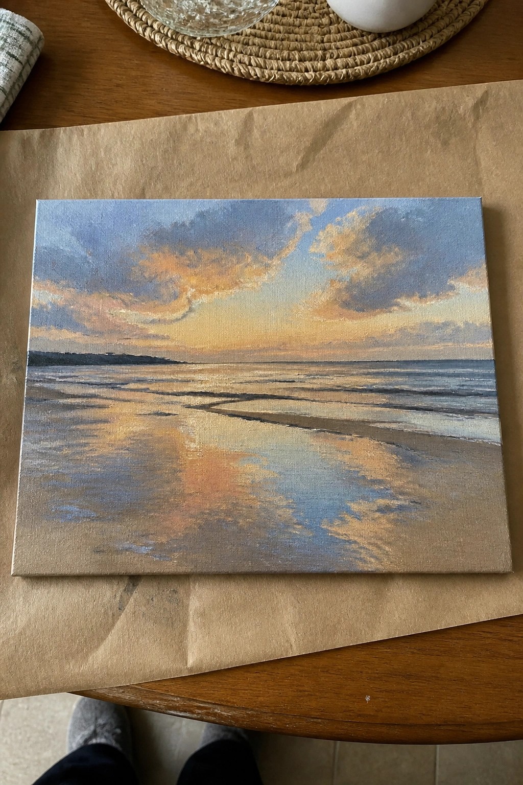 Sunset seascape painting on canvas resting on brown paper atop wooden table