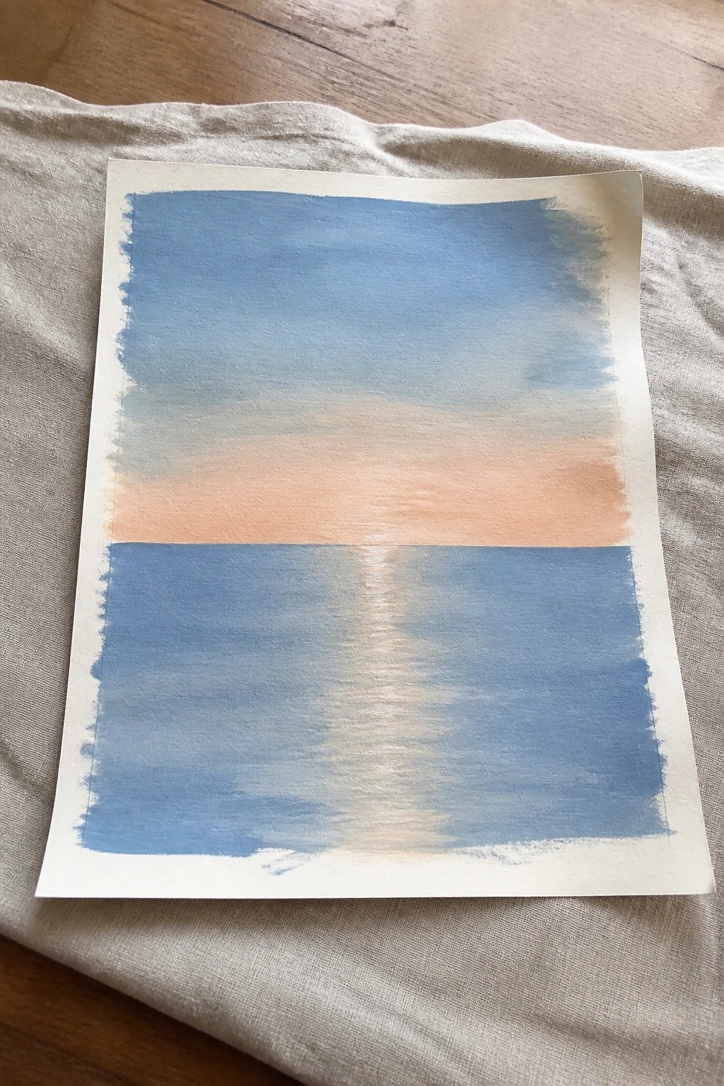 Watercolor painting of blue sky fading to peach sunset over reflective water on fabric.