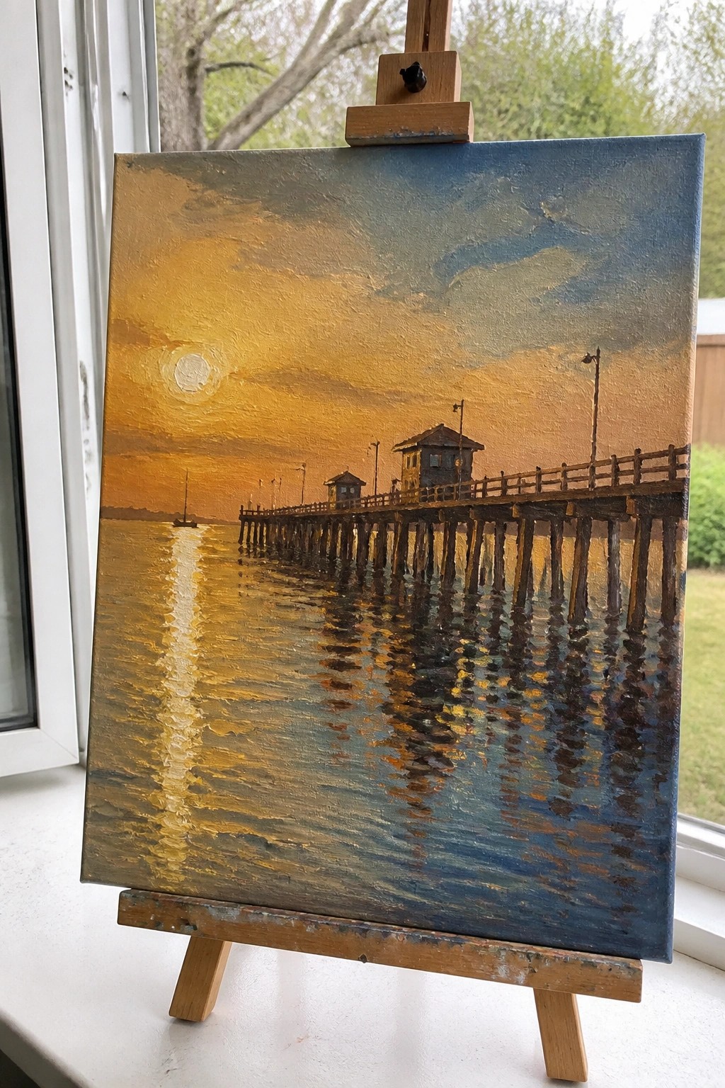 Oil painting on easel of wooden pier at golden sunset with water reflections.