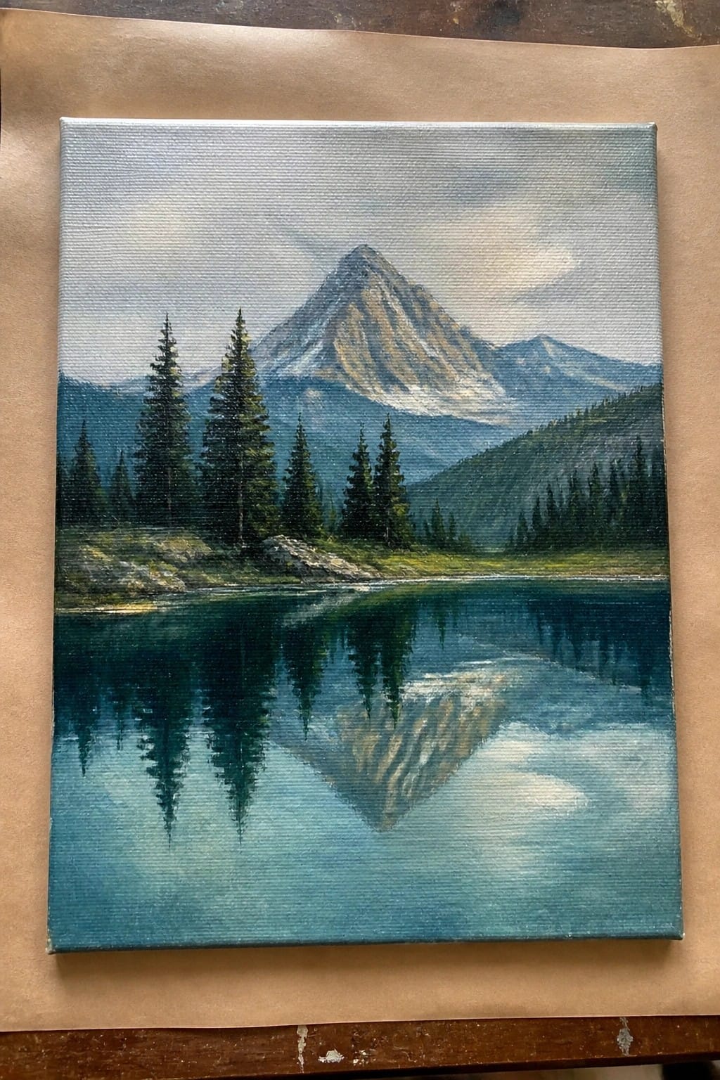 Oil painting of snow-capped mountain reflected in calm blue lake with pine trees.