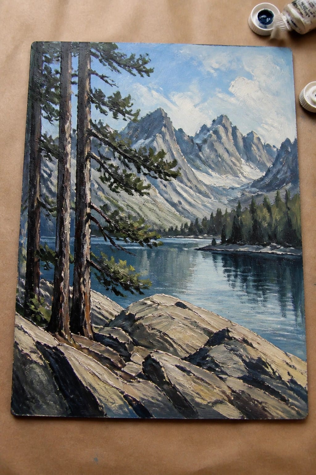 Oil painting of pine trees by rocky lake with snow-capped mountains and paint tubes nearby.