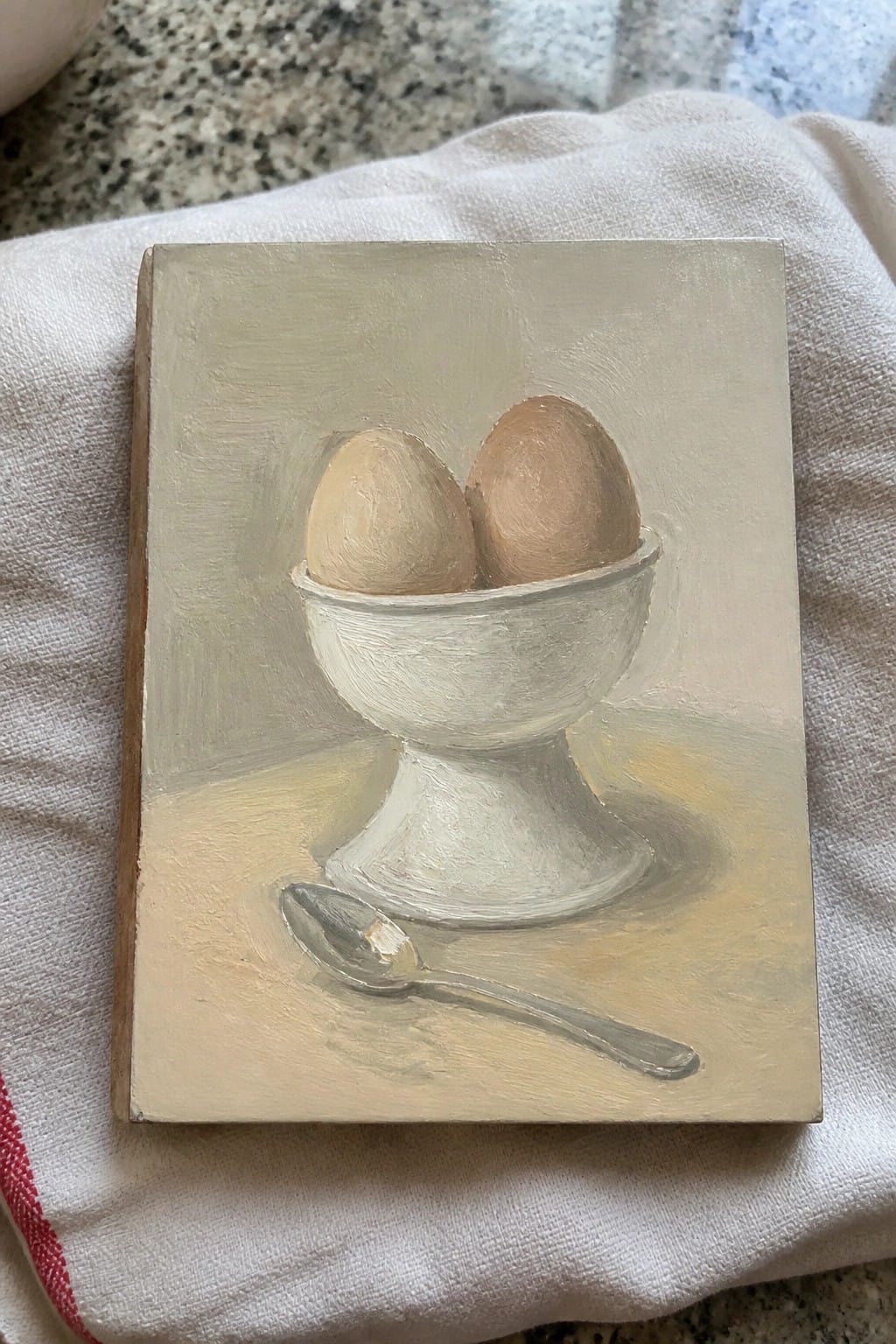 Painting of two eggs in white pedestal cup beside a spoon on cloth