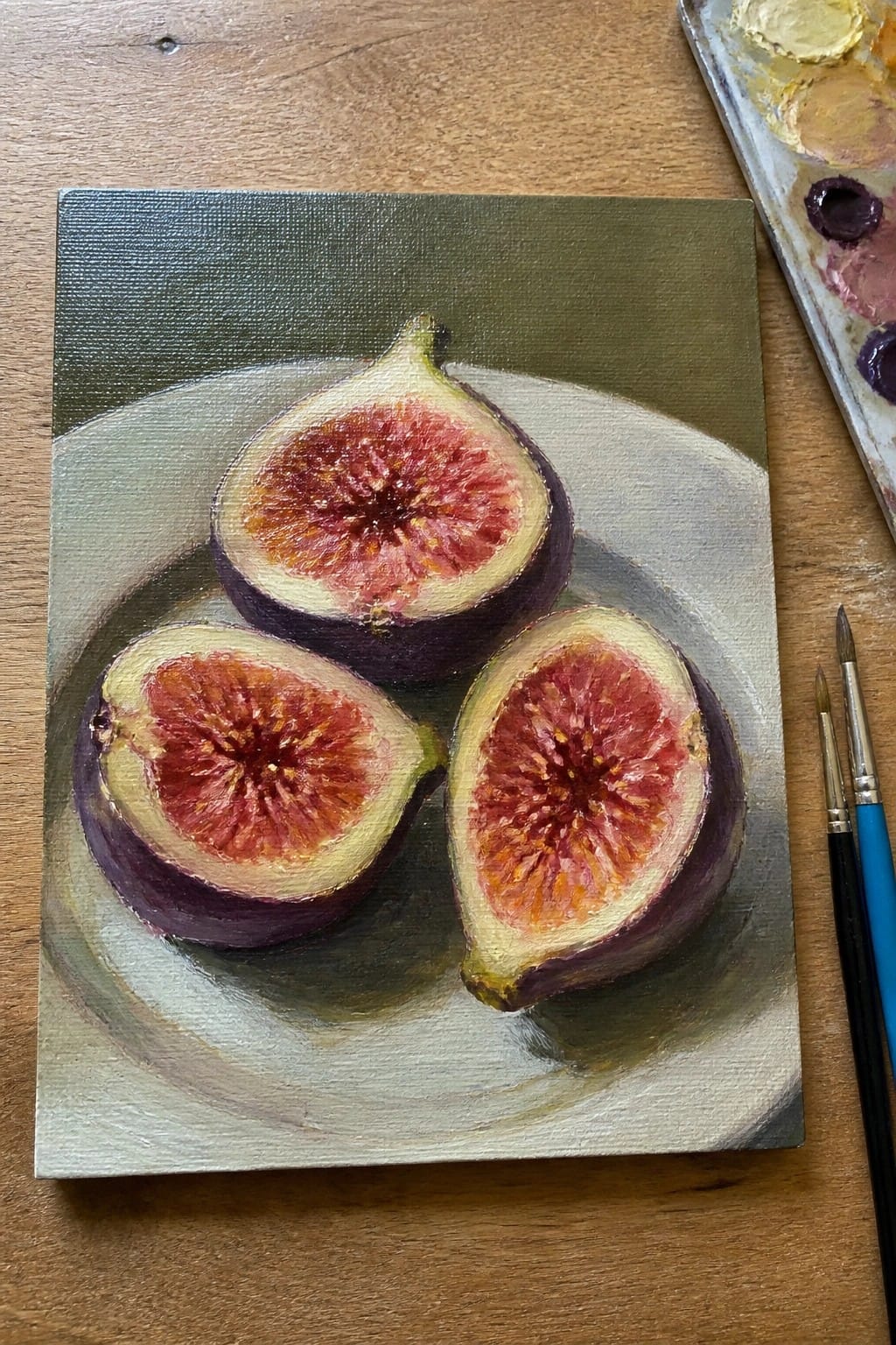 Realistic oil painting of three halved figs on a plate with palette and brushes nearby