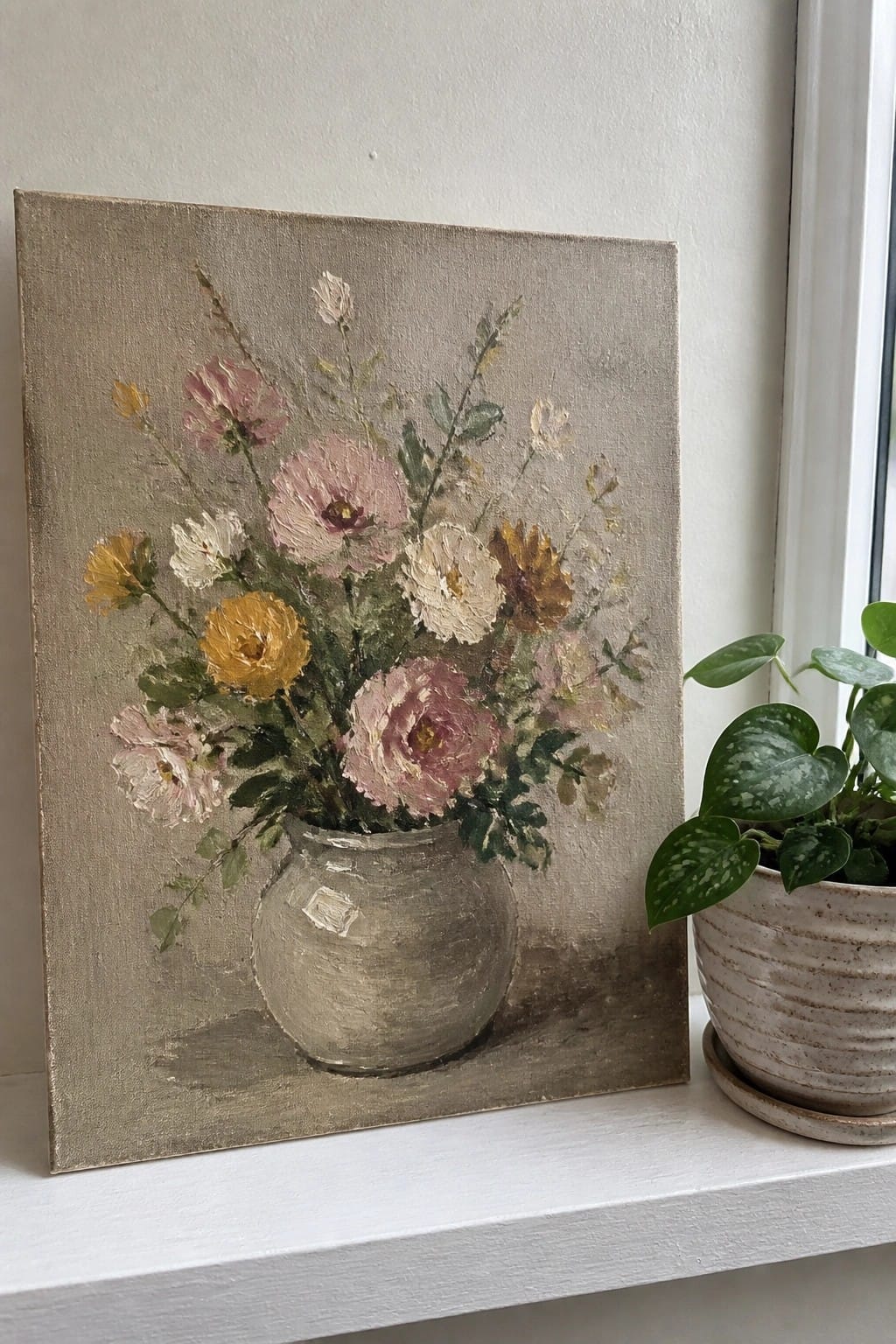 Painting of pink and yellow flowers in gray vase beside green potted plant on windowsill