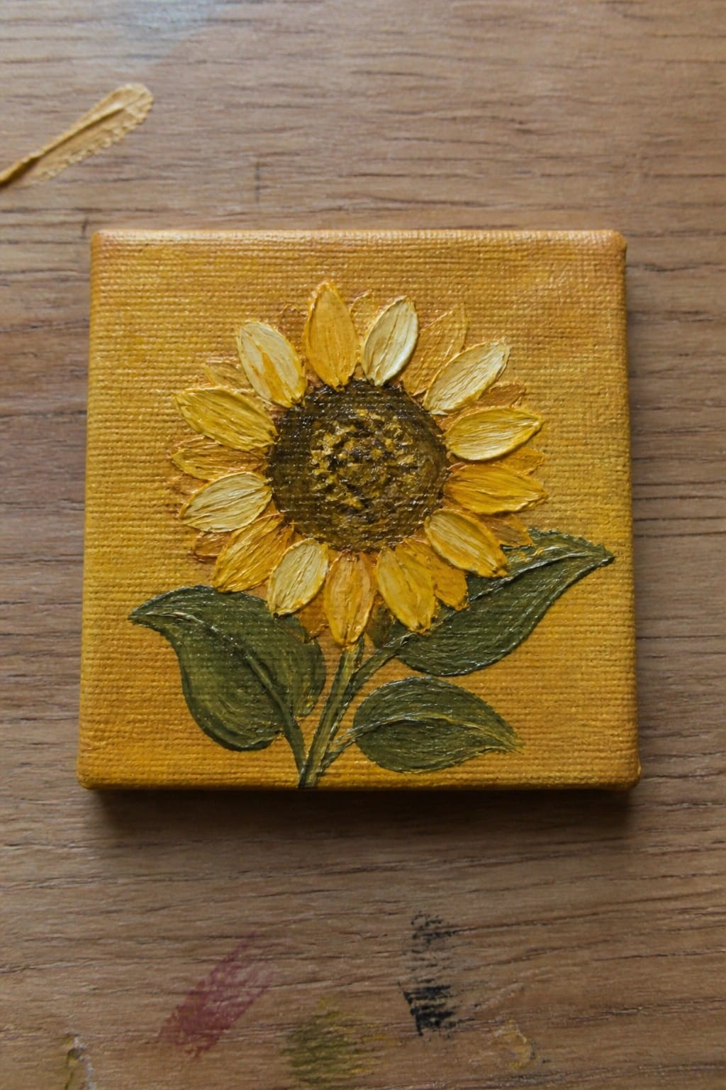 Small yellow canvas with painted sunflower, yellow petals, brown center and green leaves on wood.
