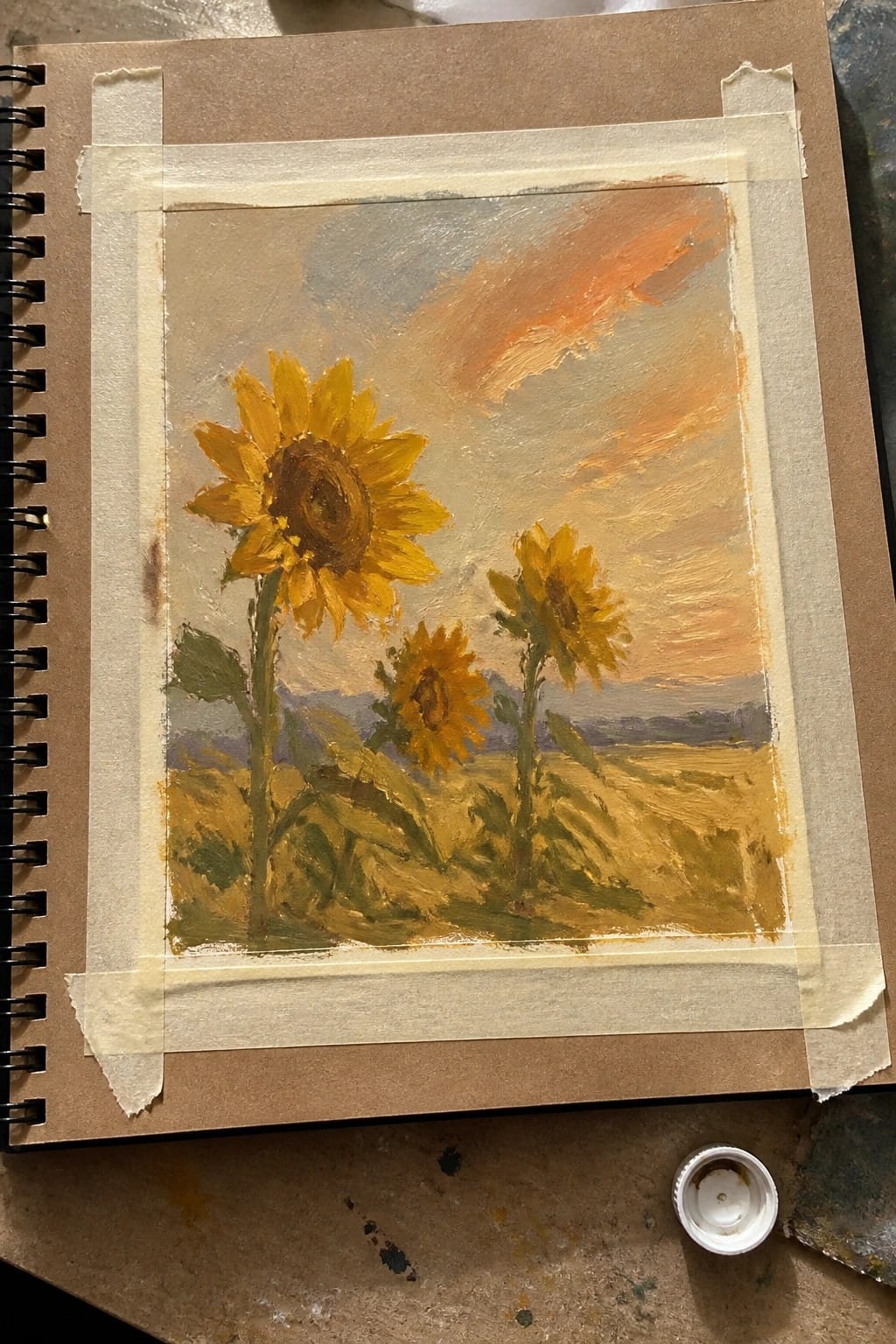 Oil painting of sunflowers in a field with a warm orange and gray sky, taped onto a sketchbook page.