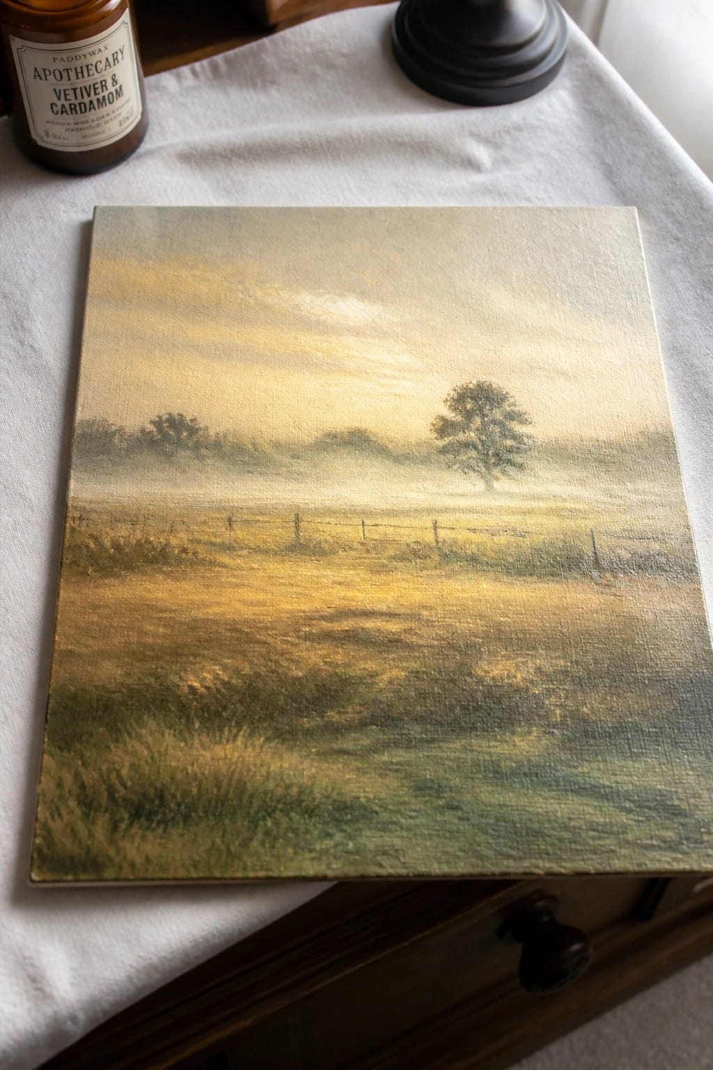 Painting of misty golden field with fence and trees beside candle jar on white cloth