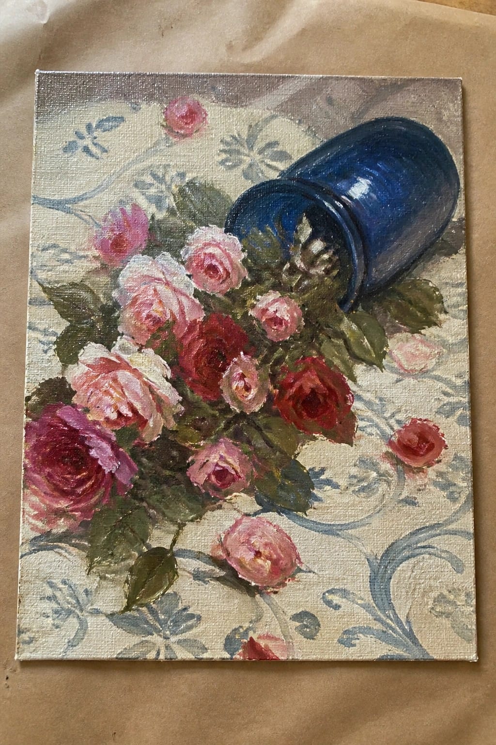An oil painting of a blue vase tipped over with pink and red roses spilling out onto patterned fabric.