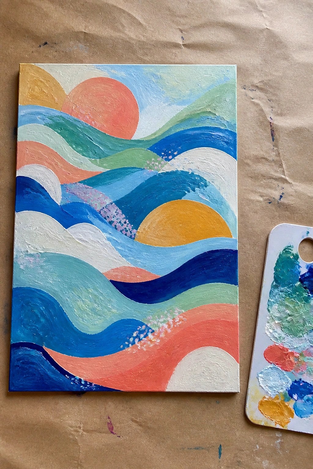 Abstract wave painting on canvas beside paint palette on brown paper