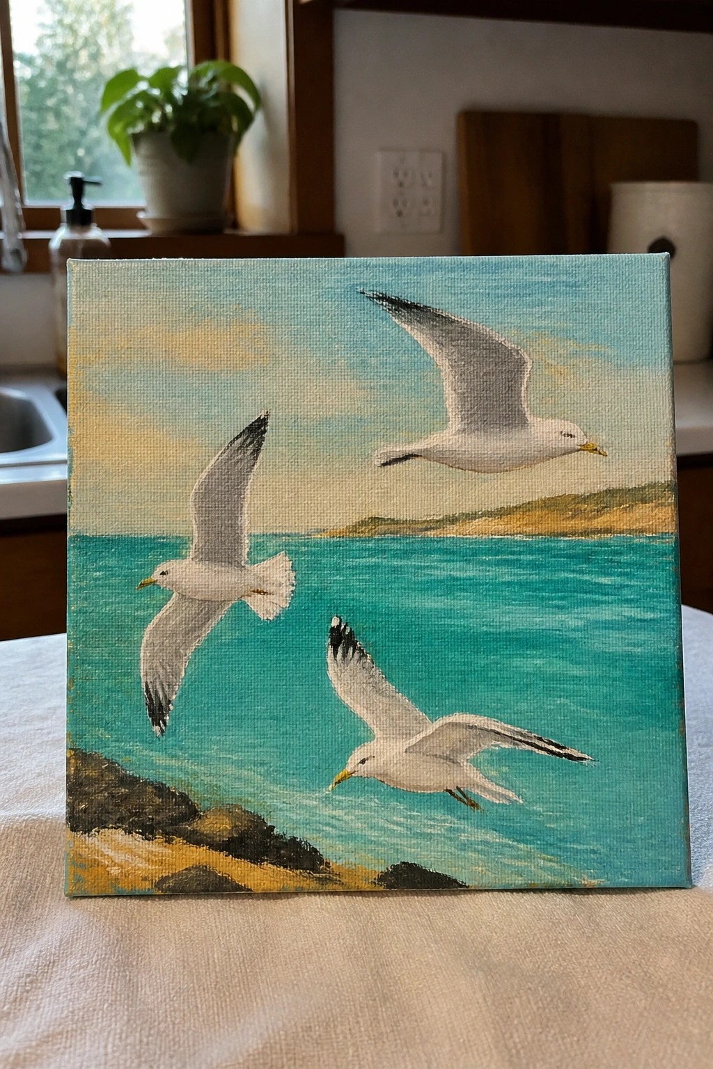 Three seagulls soar over turquoise sea near rocky shore with distant hills in painting.