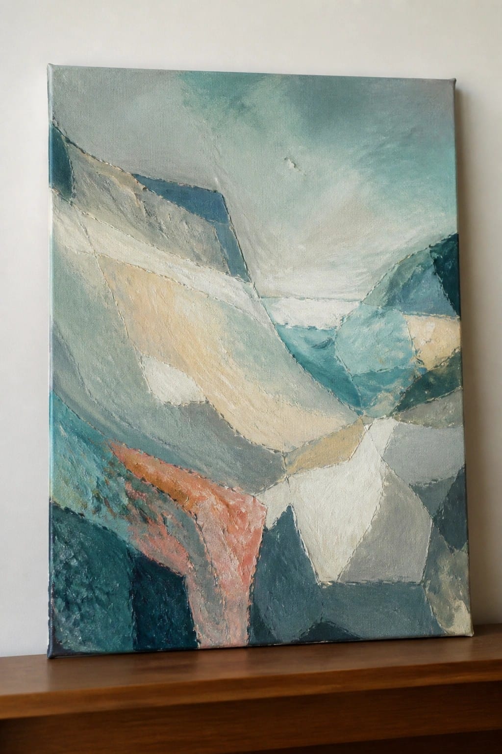 An abstract oil painting with angular teal, blue, white, and coral shapes on a canvas resting on a wooden surface.