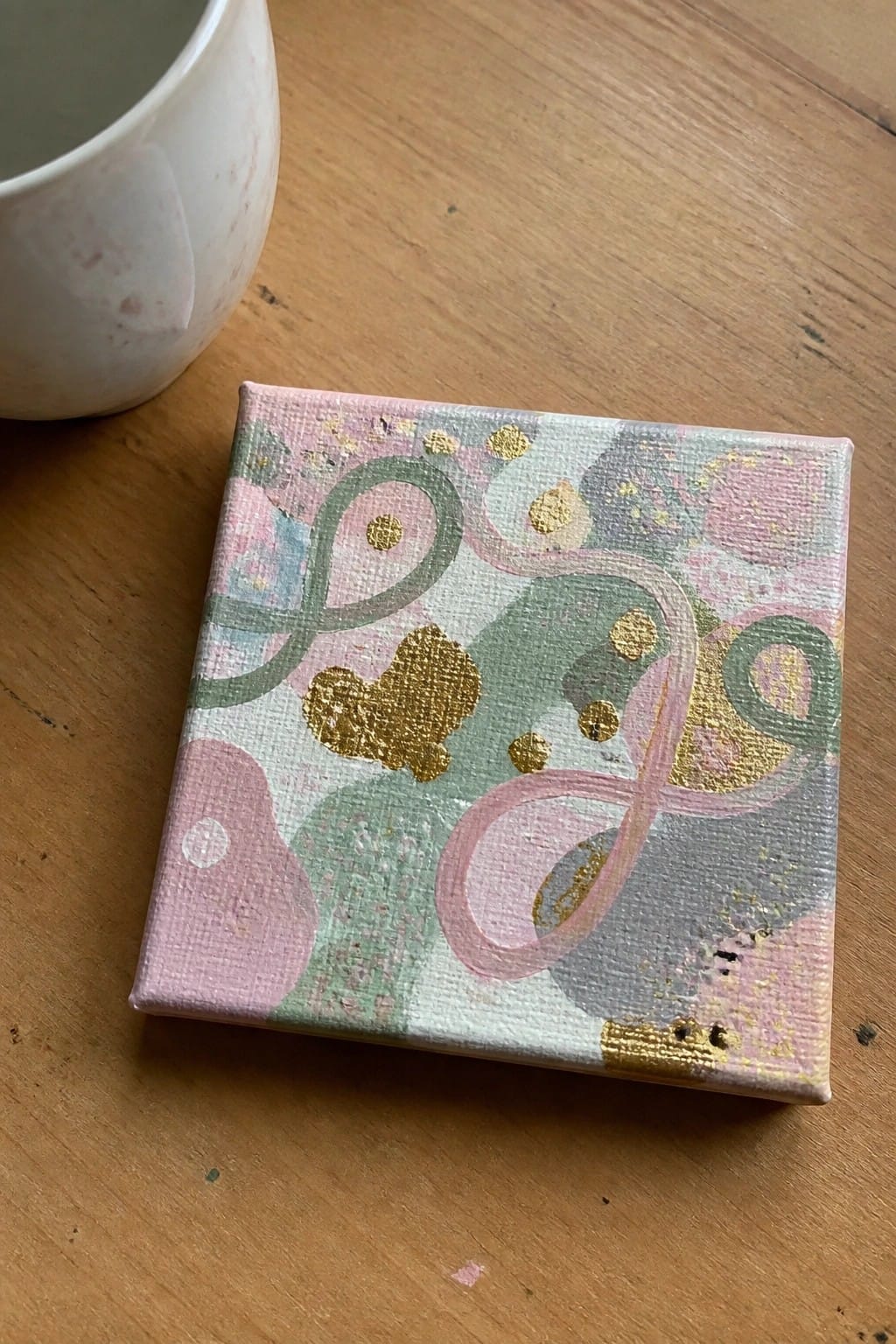 Abstract pink green gray canvas with gold accents beside white mug on wood.
