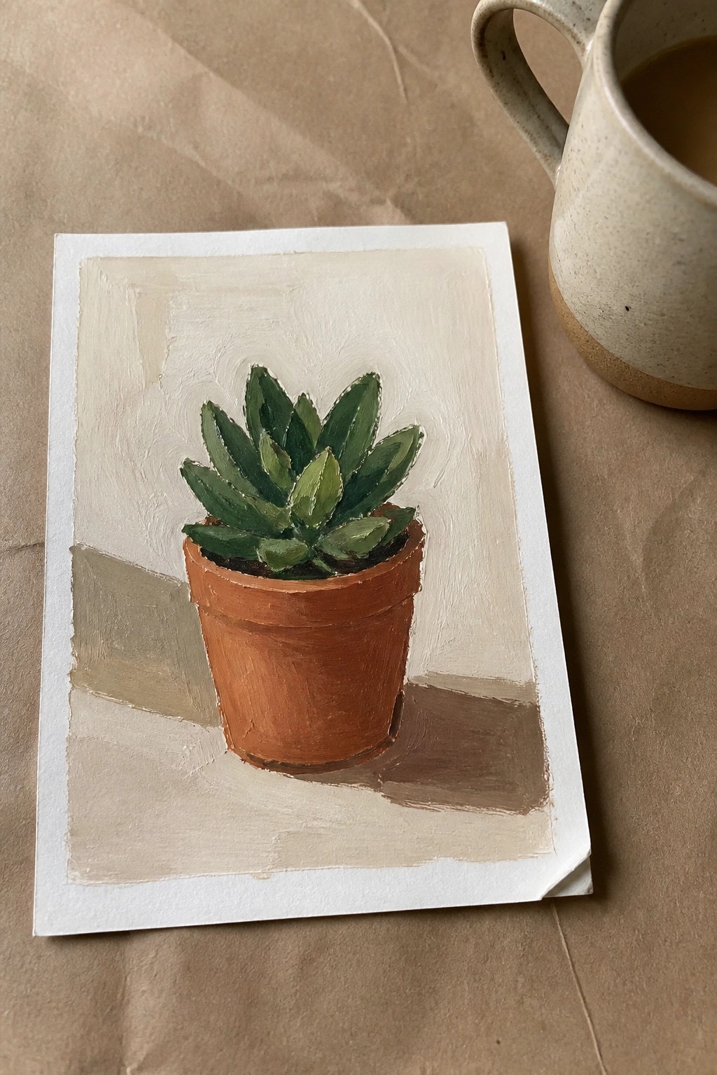 Painting of green succulent in terracotta pot beside speckled ceramic mug on brown paper