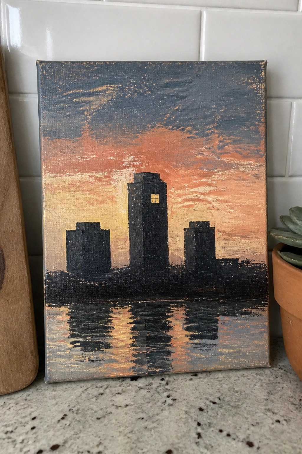 Painted canvas of silhouetted buildings against orange sunset sky with water reflections and one lit window