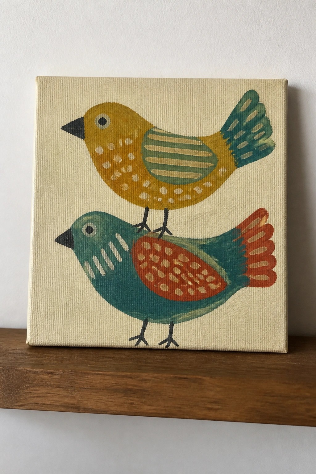 Two stacked colorful birds painted on square canvas atop wooden shelf.