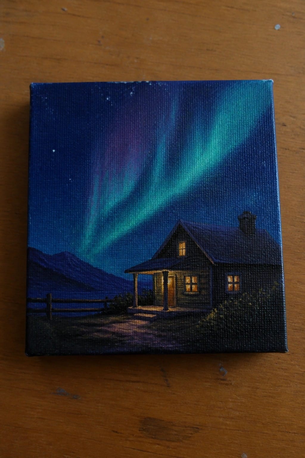Small canvas painting of a lit wooden cabin under vibrant green-purple northern lights.