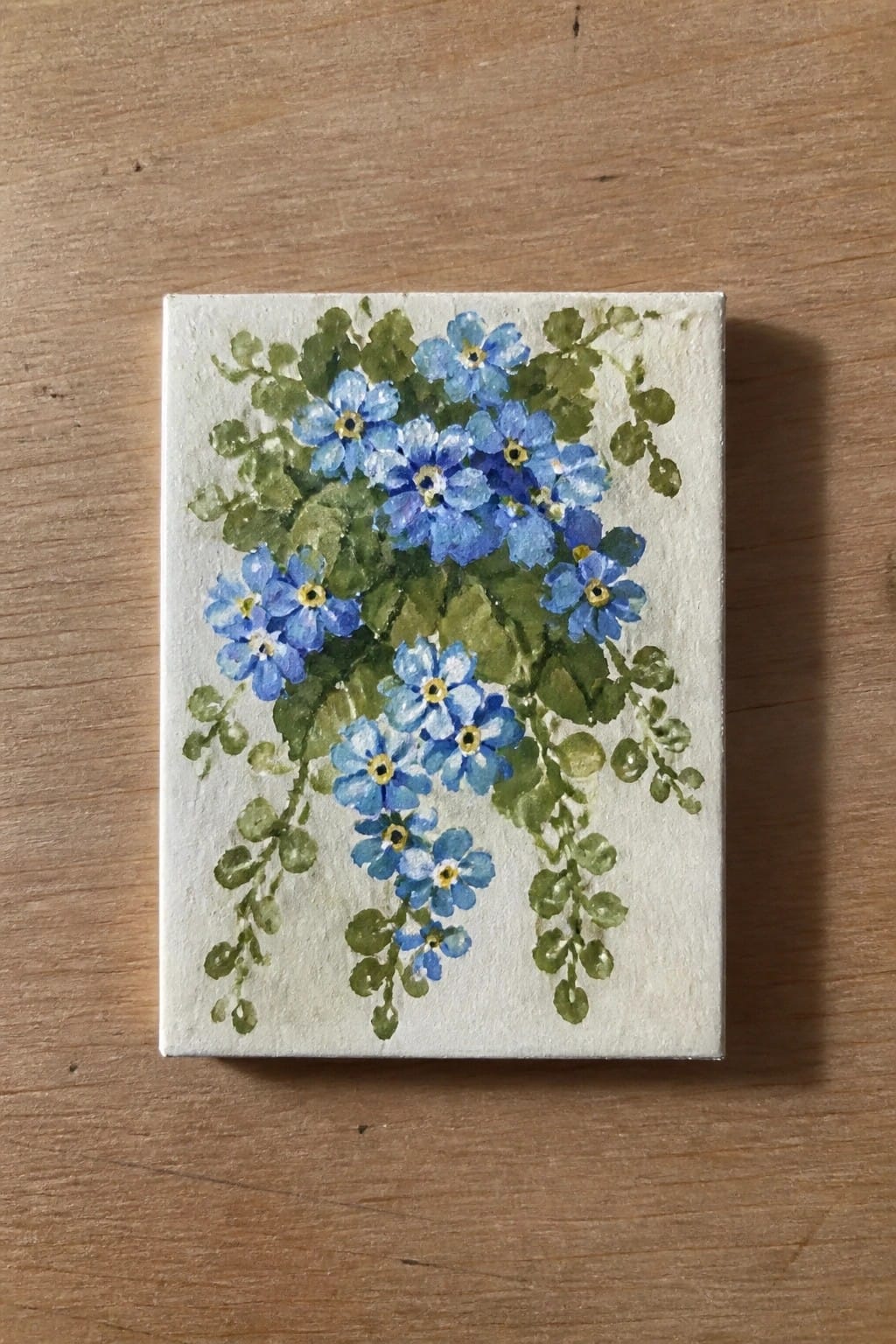 Watercolor painting of blue flowers with yellow centers and green trailing leaves on canvas