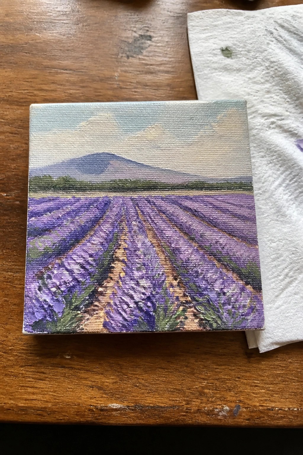 Small canvas painting of lavender fields with mountains beside paint-stained paper towel on wood table