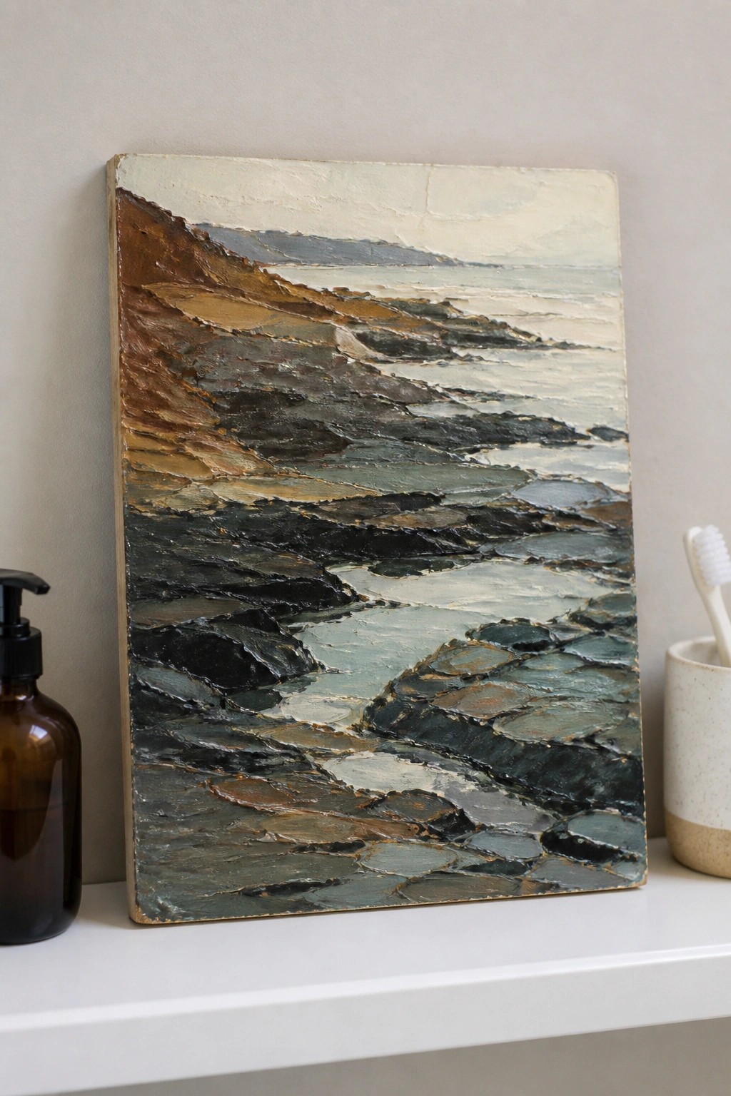 Thickly textured coastal landscape painting on shelf beside pump bottle and toothbrush holder