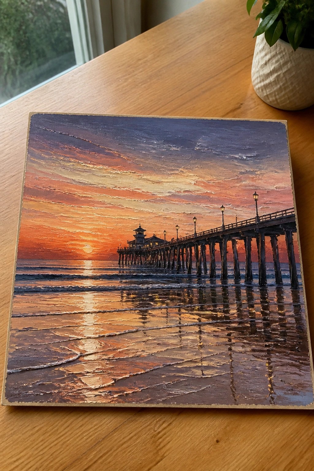 Textured painting of a long pier at vibrant orange ocean sunset on wood table