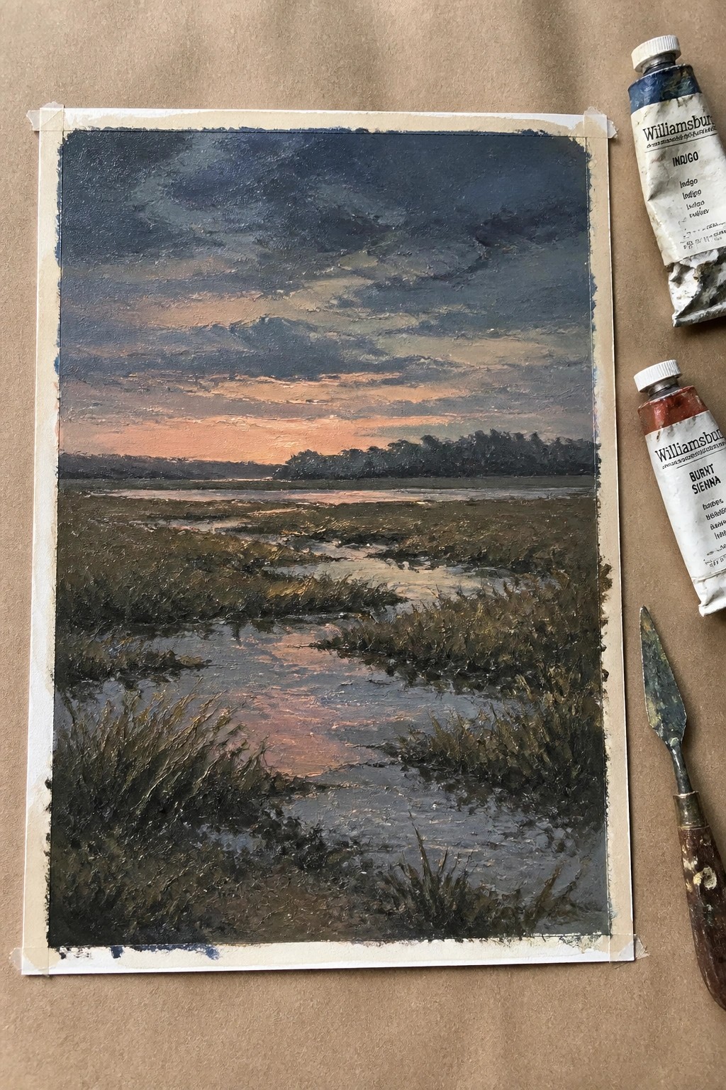 Oil painting of a marsh at dusk with a reflective stream, dark clouds, orange horizon, and textured grassy banks next to indigo and burnt sienna paint tubes and a palette knife.