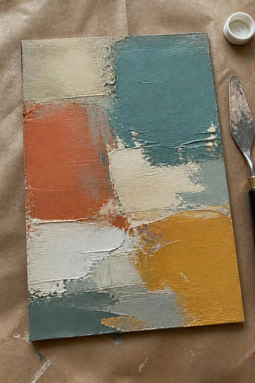 Abstract painting with textured teal, orange, yellow, white and beige blocks beside palette knife.