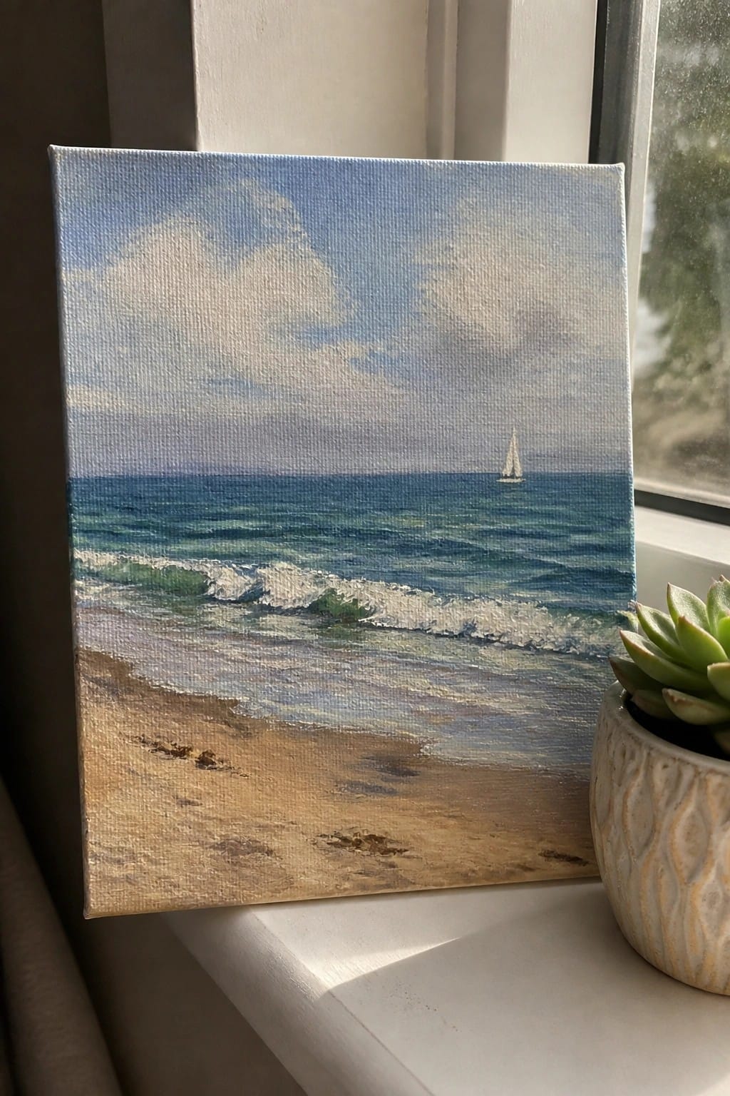 A small oil painting of a beach scene with waves, ocean, sky, and a sailboat resting on a white windowsill next to a potted succulent.
