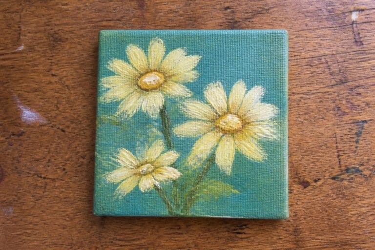 19 Essential Oil Painting Setup Ideas For Beginners In Small Spaces Small square painting of three yellow daisies on green background atop wood.