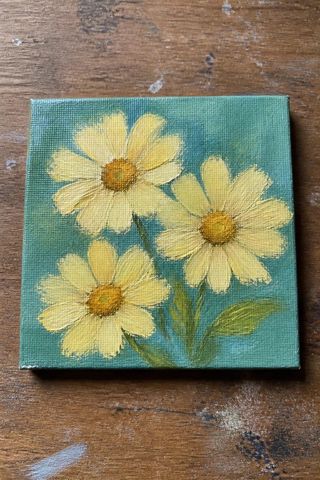 Small square painting of three yellow daisies on green background atop wood.