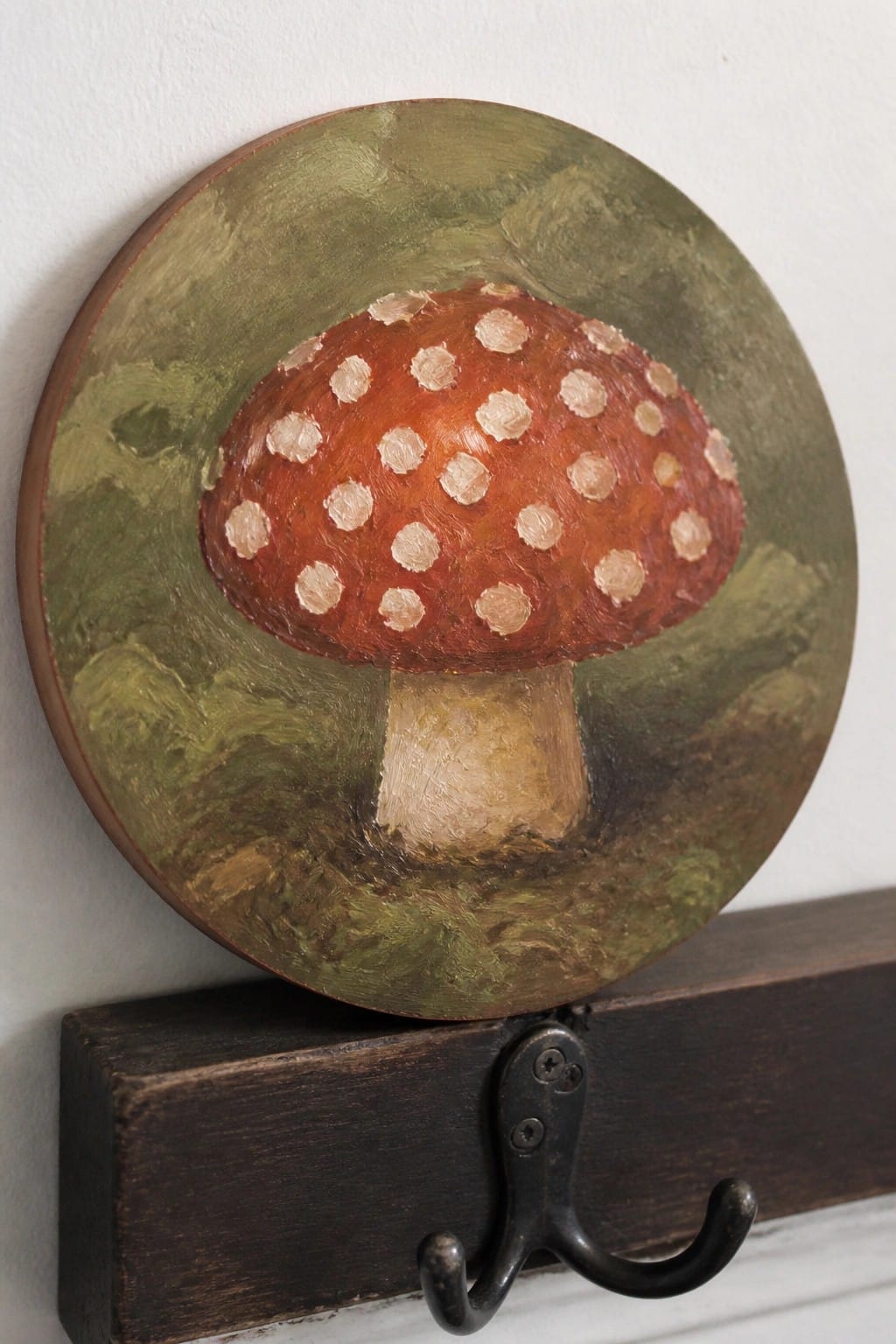 Circular red mushroom painting on green plaque above black metal hook on wood.