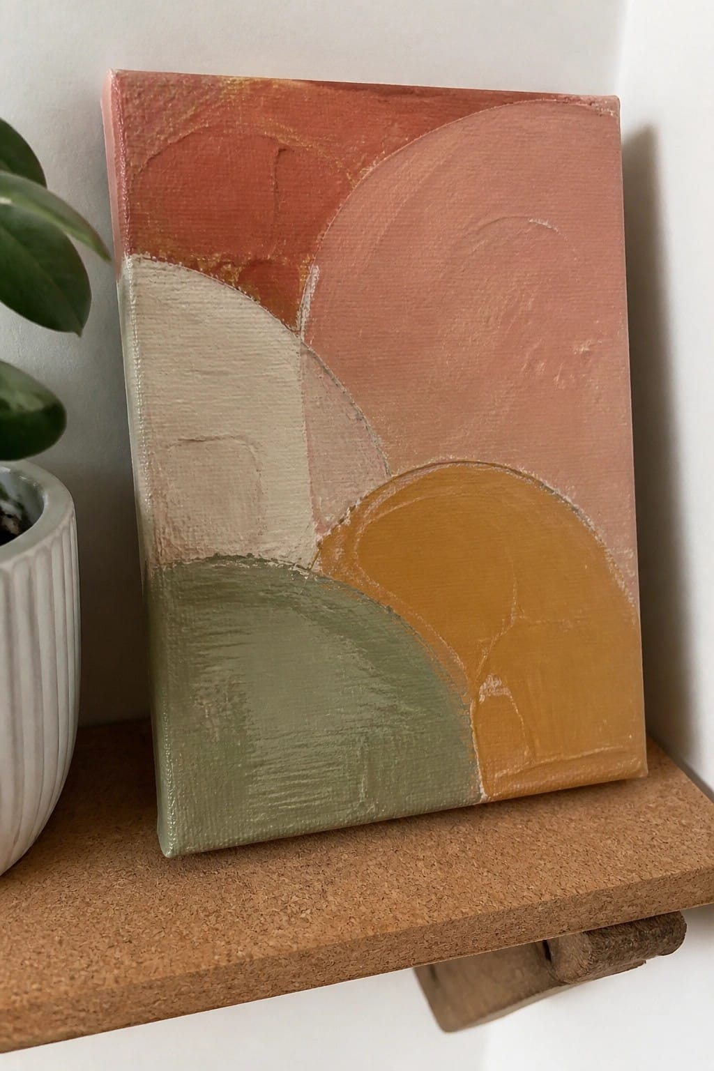 Abstract painting with curved shapes in terracotta, peach, green and mustard on wooden shelf beside plant