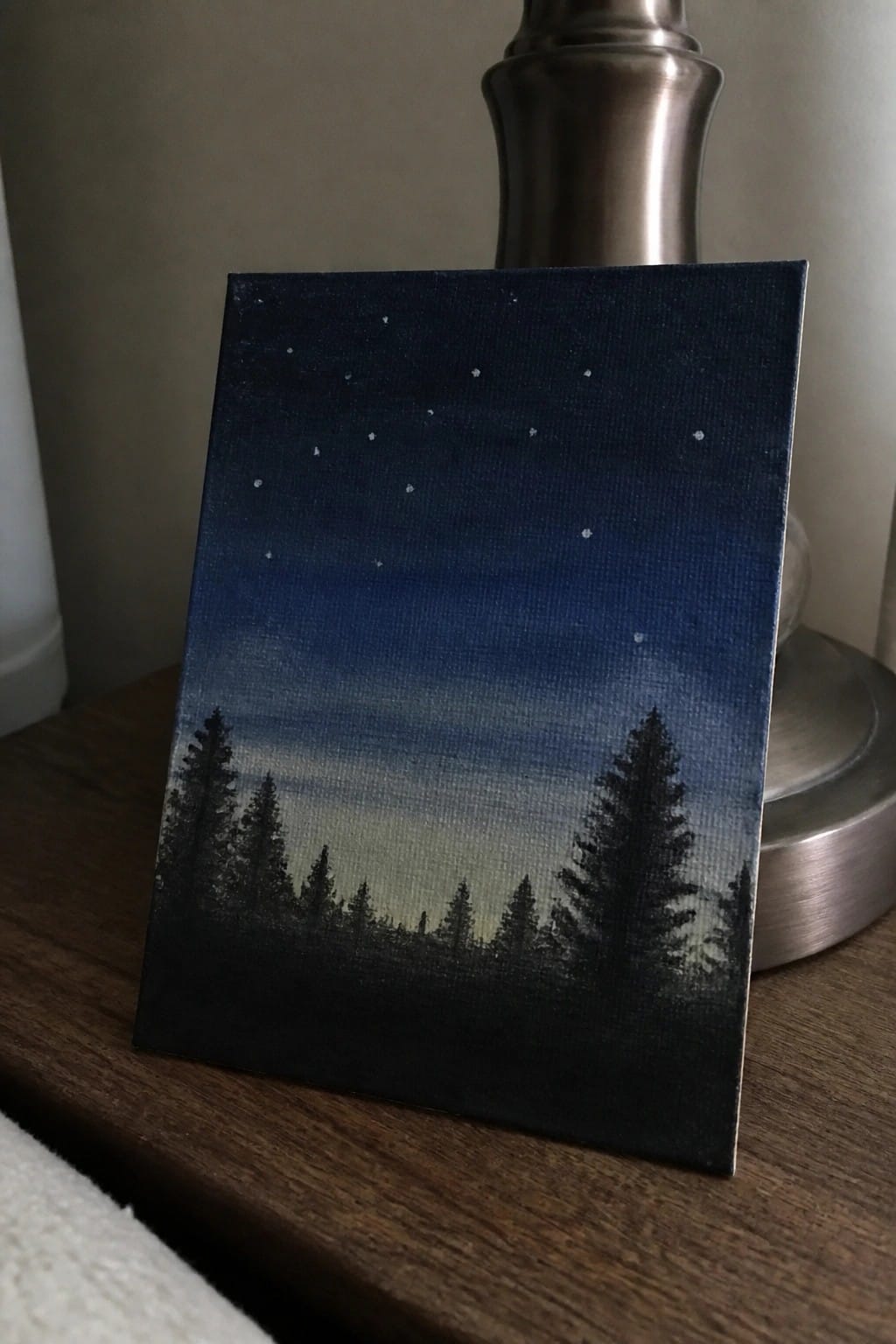 Starry night sky painting with pine tree silhouettes on wooden table beside lamp base