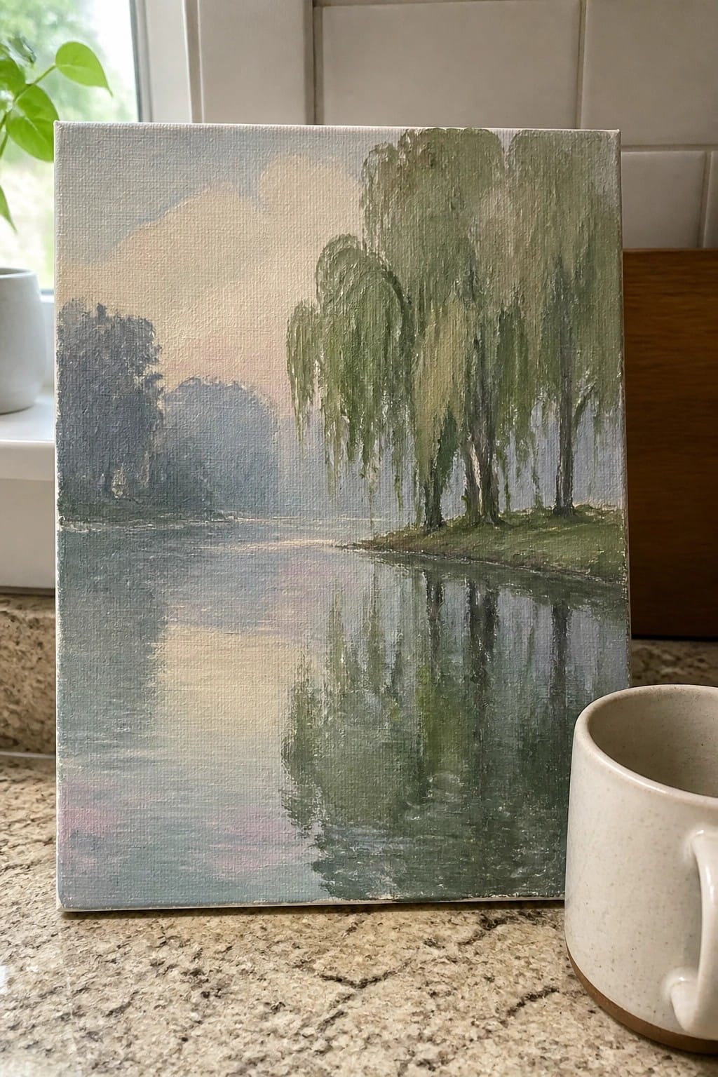 Landscape painting of weeping willows reflected in misty river, beside speckled mug.