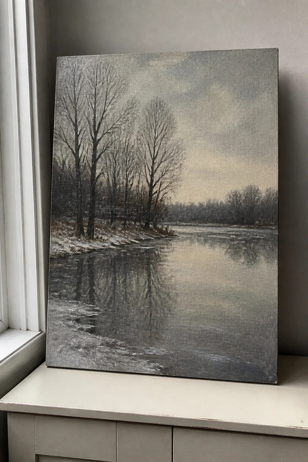 Monochrome painting of snowy river with bare trees propped beside window.